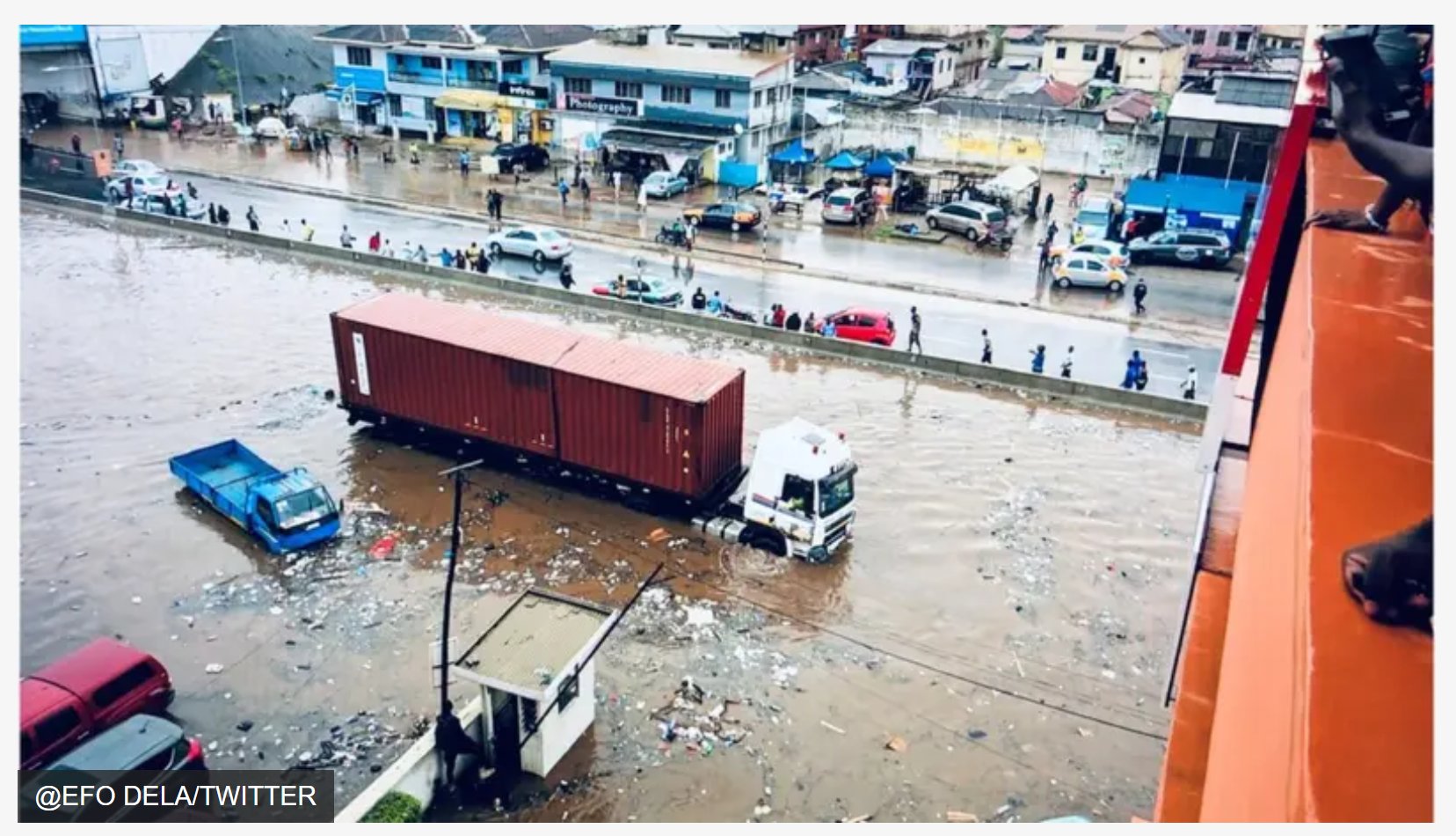 A stranded articulated truck and scattered debris in floodwater on an Accra street, as onlookers watch from higher ground